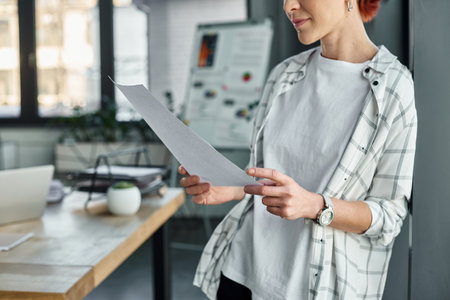 cropped view of non-binary person in casual attire standing with paper documents in modern officeの写真素材