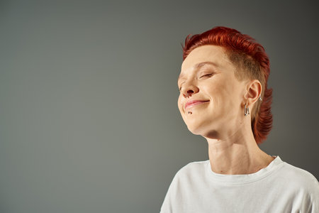 portrait of redhead bigender person with facial piercing smiling with closed eyes on grey backdrop