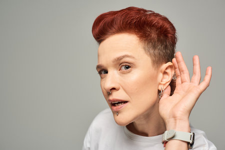 curious redhead bigender person holding hand near ear while looking at camera on grey backdrop