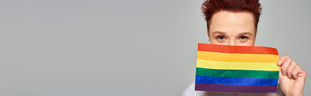 joyful redhead queer person obscuring face with small LGBT flag looking at camera on grey, banner