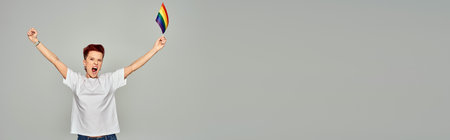 excited queer person in white t-shirt standing with small LGBT flag and screaming on grey, bannerの写真素材