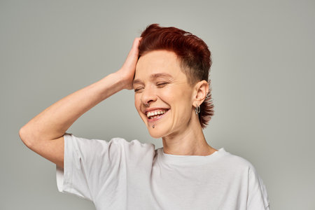 cheerful non-binary person in white t-shirt touching red hair and smiling with closed eyes on greyの写真素材