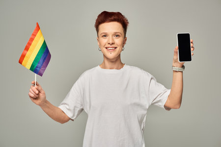 joyful redhead queer person holding small LGBT flag and smartphone with blank screen on greyの写真素材