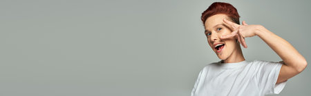 excited redhead bigender person in white t-shirt showing victory sign near face on grey, bannerの写真素材