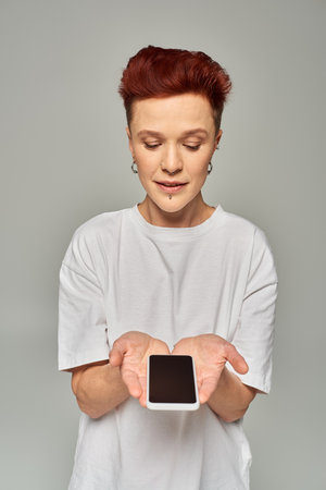 redhead non-binary person in white t-shirt holding mobile phone with blank screen on grey backdrop