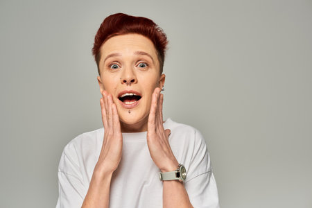 amazed redhead queer person with open mouth touching face and looking at camera on grey backdropの写真素材