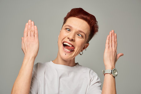 funny redhead bigender person in white t-shirt grimacing and sticking out tongue on grey backdropの写真素材