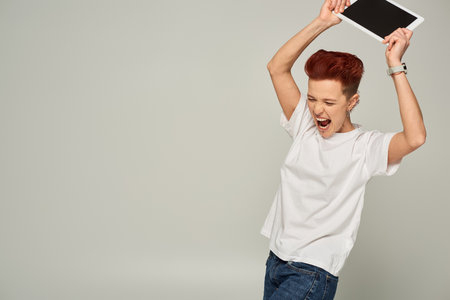 angry queer person in white t-shirt screaming and breaking digital tablet with blank screen on grey