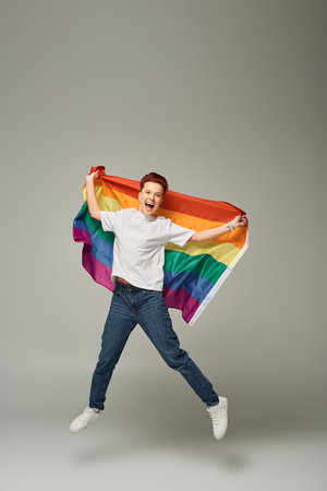 overjoyed queer person in white t-shirt and jeans jumping and levitating with LGBT flag on greyの写真素材