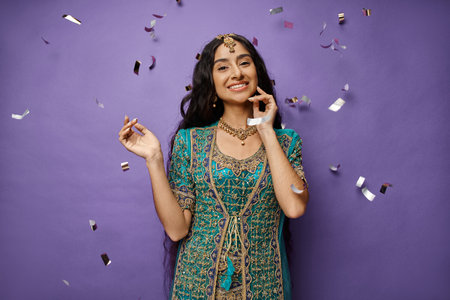cheerful young indian woman with long hair smiling at camera while posing under confetti rainの写真素材