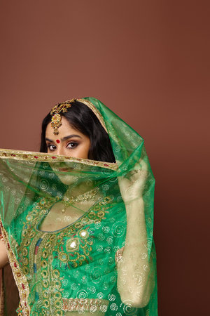 vertical shot of indian young woman covering her face with green veil posing on brown backdropの写真素材