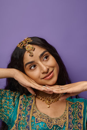 vertical shot of beautiful joyful indian woman posing with hands under chin and looking away