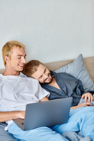 smiling gay couple watching romantic movie on laptop in bedroom, happy morning leisure on bedの写真素材