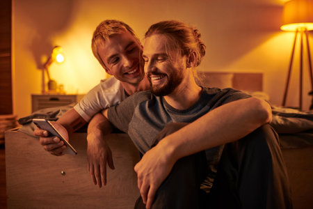 joyful bearded gay man browsing internet on mobile phone near smiling boyfriend in bedroom at nightの写真素材