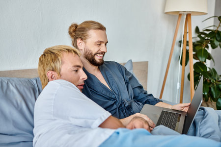 joyful bearded gay man smiling while watching comedy movie on laptop with boyfriend in bedroomの写真素材