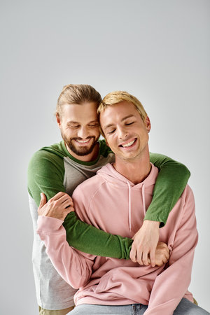 joyful bearded gay man embracing stylish boyfriend smiling with closed eyes on grey backdropの写真素材