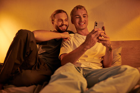 smiling gay man browsing social media on smartphone near happy boyfriend in bedroom at nightの写真素材