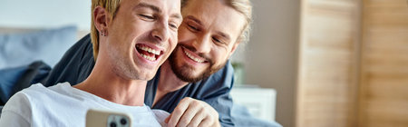 cheerful tattooed gay man browsing internet on smartphone near smiling boyfriend in bedroom, bannerの写真素材