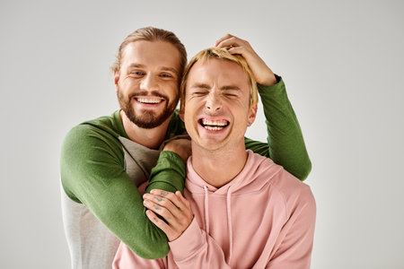 cheerful bearded gay man embracing trendy boyfriend while having fun on grey backdrop in studioの写真素材