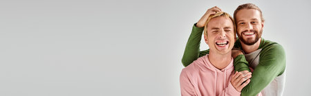 cheerful bearded gay man embracing trendy boyfriend while having fun on grey backdrop, bannerの写真素材