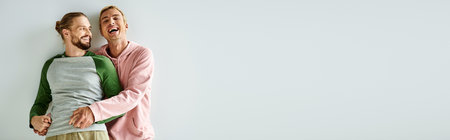 excited gay man holding hands of bearded boyfriend and laughing on grey backdrop, horizontal bannerの写真素材