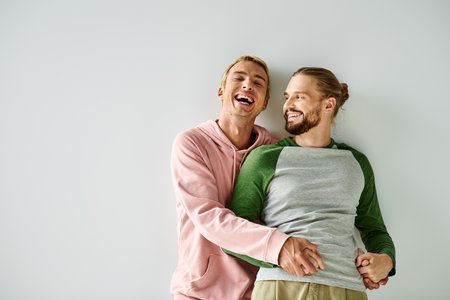 excited gay man holding hands of bearded boyfriend and laughing on grey backdrop, same-sex coupleの写真素材