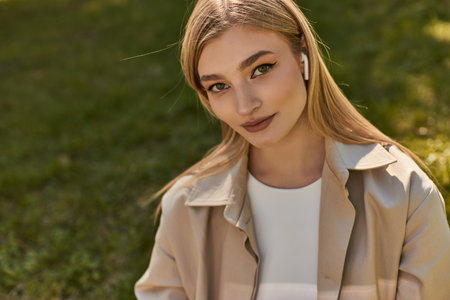 portrait of pretty young woman in wireless earphones and beige trench coat looking at camera in parkの写真素材