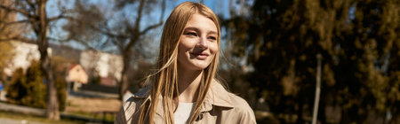 happy blonde woman in stylish beige trench coat smiling while walking in park, horizontal banner