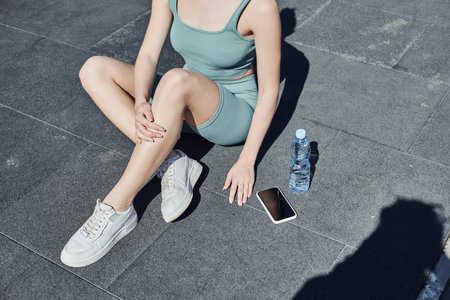 cropped sportswoman in tight activewear sitting next to bottle of water and smartphone on floorの写真素材