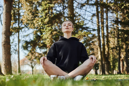 pretty young woman with blonde hair and sportswear sitting on mat while meditating in parkの写真素材