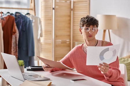 curly androgynous freelancer in pink sweater and eyeglasses working remotely near devices on deskの写真素材