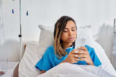 young african american woman lying in hospital bed and looking at her mobile phone, healthcareの写真素材