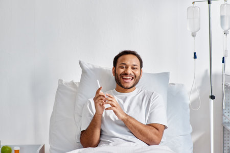 joyful indian man lying in hospital bed with phone in hands and smiling cheerfully, healthcareの写真素材