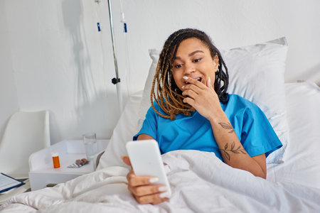 attractive young african american woman having video call while lying in hospital bed, healthcareの写真素材