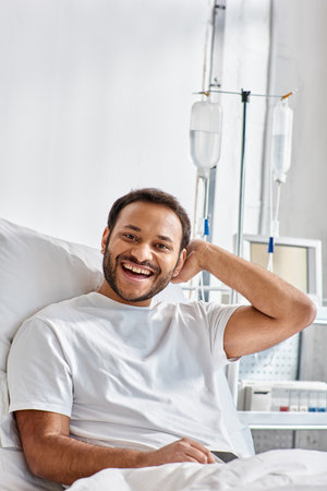 joyous young indian man lying in hospital bed and smiling cheerfully at camera, healthcareの写真素材