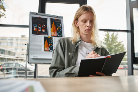 young focused non binary person in green shirt taking notes sitting at desk, business concept
