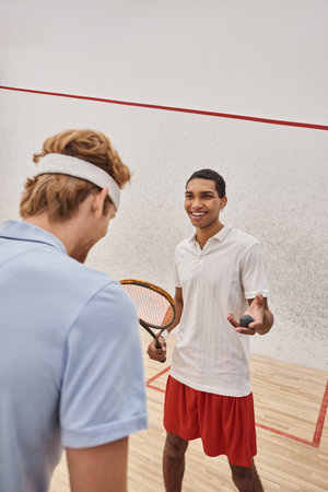 cheerful african american man with squash ball and racquet standing near friend inside of courtの写真素材