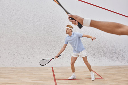 redhead man in sportswear and headband playing with african american friend inside of squash courtの写真素材