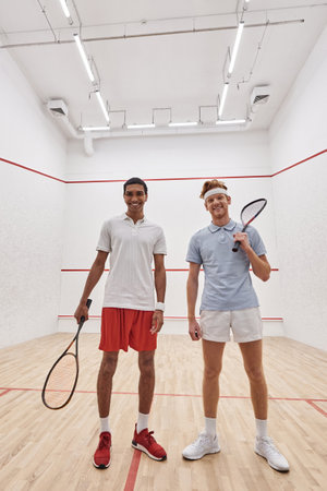 vertical shot, happy interracial male friends looking at camera and holding racquets for squashの写真素材