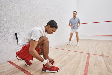 african american man tying shoelaces while sitting inside of squash court near redhead friendの写真素材
