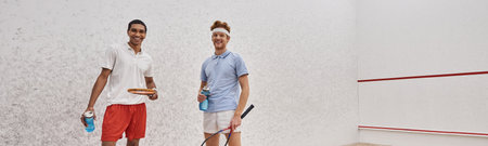 cheerful interracial sportsmen with squash racquets and bottles with water standing in court, bannerの写真素材