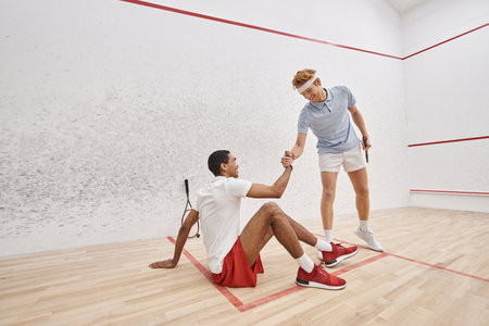 happy redhead man helping his african american friend to stand up from floor inside of squash courtの写真素材