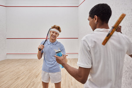 joyful interracial sportsmen with squash racquets and bottles with water chatting  in courtの写真素材