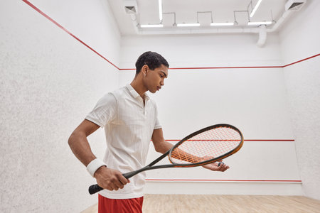 african american man in active wear holding squash ball and racquet while playing inside of courtの写真素材