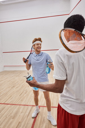 smiling interracial sportsmen with squash racquets and bottles with water chatting  in courtの写真素材