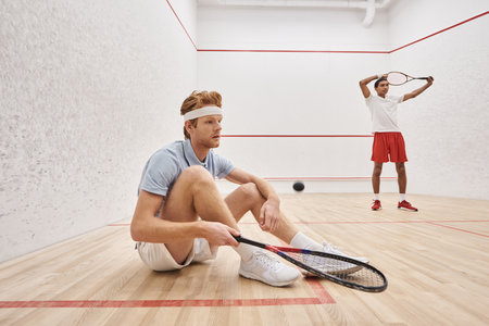man in sportswear holding racquet while sitting near african american friend inside of squash courtの写真素材