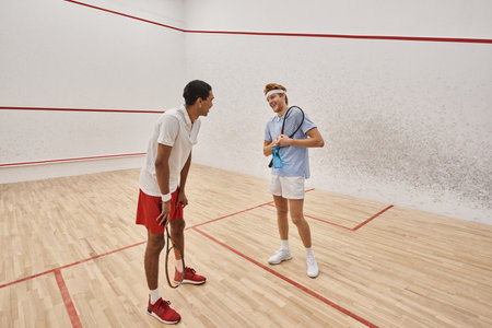 cheerful interracial male friends with squash racquets and bottles with water chatting  in courtの写真素材