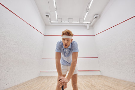 tired redhead man in headband and sportswear breathing heavily after playing squash in court
