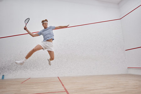motion shot, athletic sportsman holding racquet and jumping while playing squash inside of courtの写真素材
