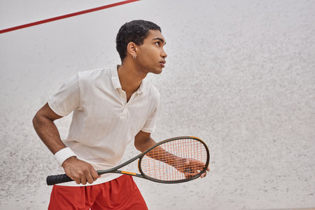 side view, athletic african american man holding squash racquet while playing game inside of courtの写真素材